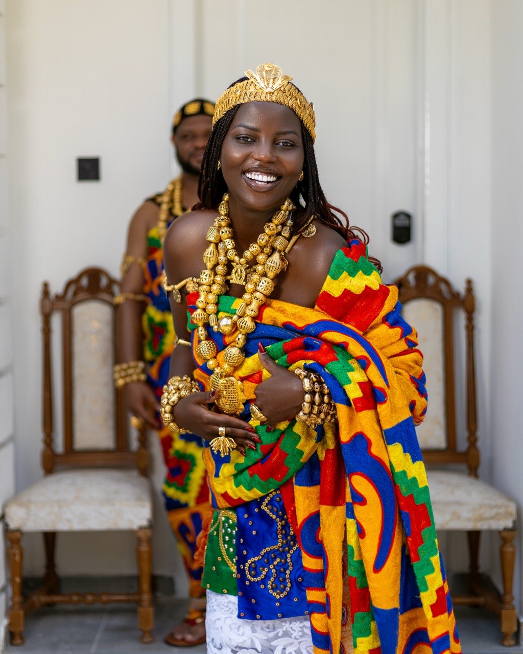 Claudine smiling in kente and gold jewellery with Edward behind her